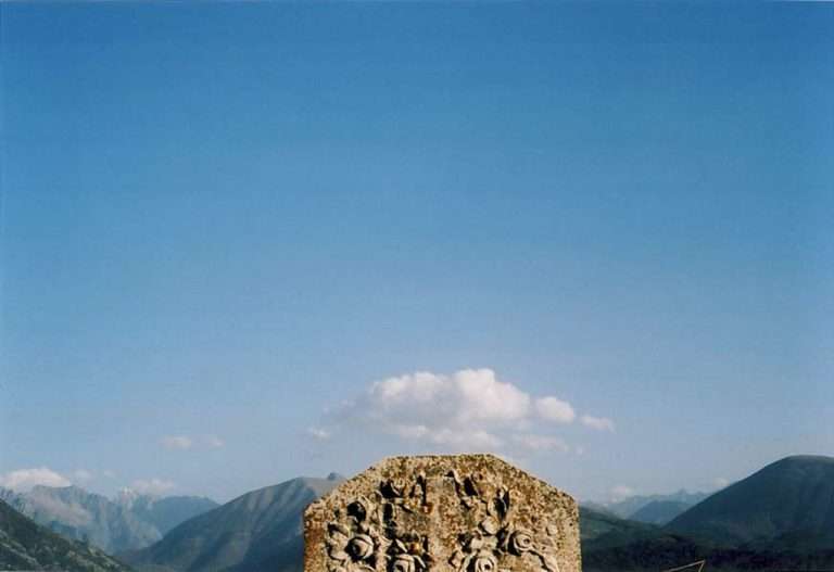 Une pierre tombale ornée d'une croix dans un paysage de montagne avec un nuage centré sur la pierre.